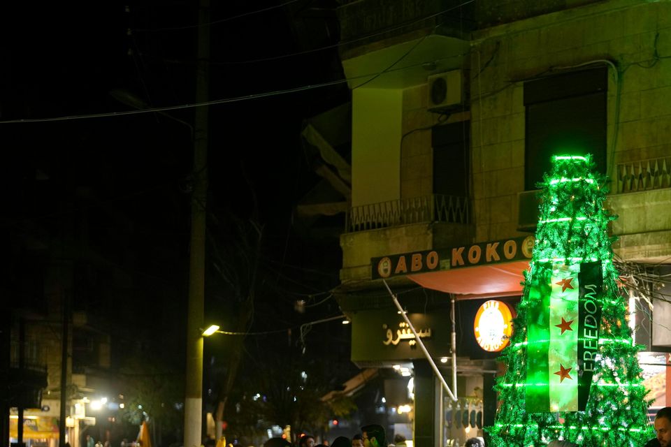 People stand next to a illuminated Christmas tree decorated with the ‘revolutionary’ Syrian flag in the city of Aleppo (Khalil Hamra/AP)