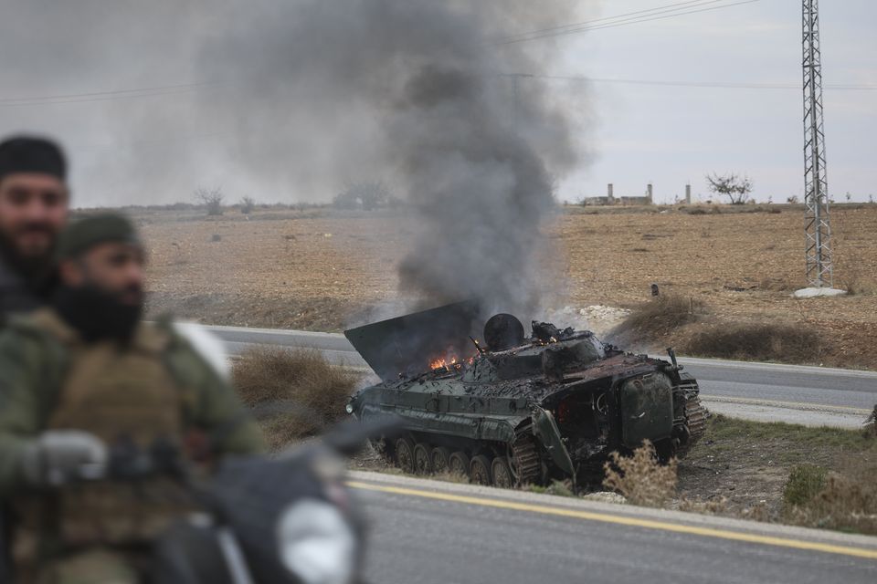 Syrian opposition fighters drive past a burning government armoured vehicle south of Hama (Ghaith Alsayed/AP)