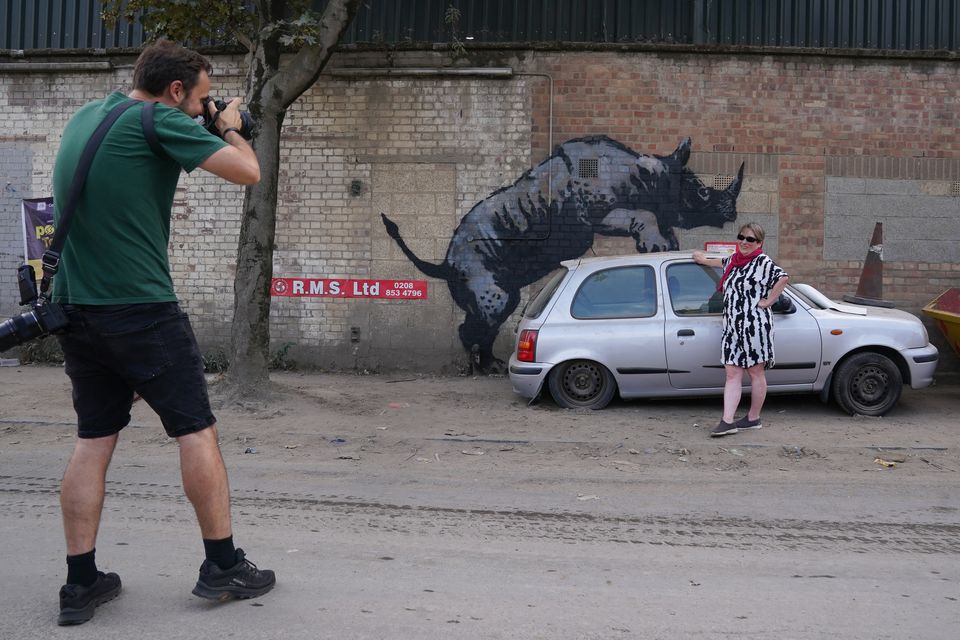 A woman poses for a photograph by a new artwork unveiled by Banksy, depicting a rhinoceros which looks as though it is climbing on top of a car (Lucy North/PA)