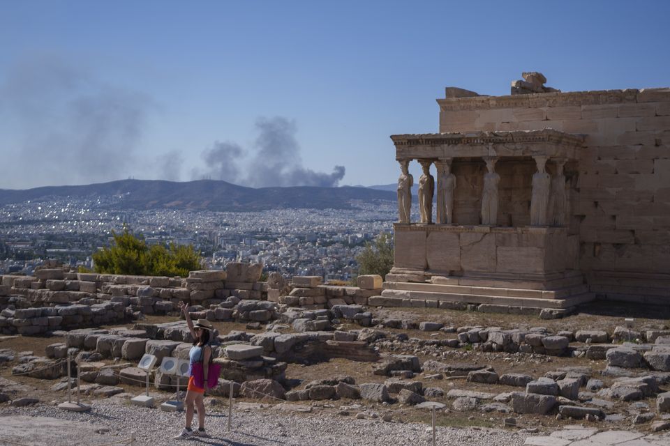 A woman poses for a photo in front Caryatid statues as smoke from a fire rises up in the background during a hot, windy day at Acropolis hill in Athens (AP Photo/Petros Giannakouris)