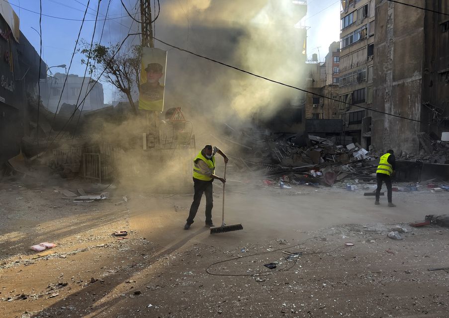 Workers clean a street as smoke rises from a destroyed building that was hit by an Israeli air strike in the southern Beirut suburb of Dahiyeh (Hussein Malla/AP)